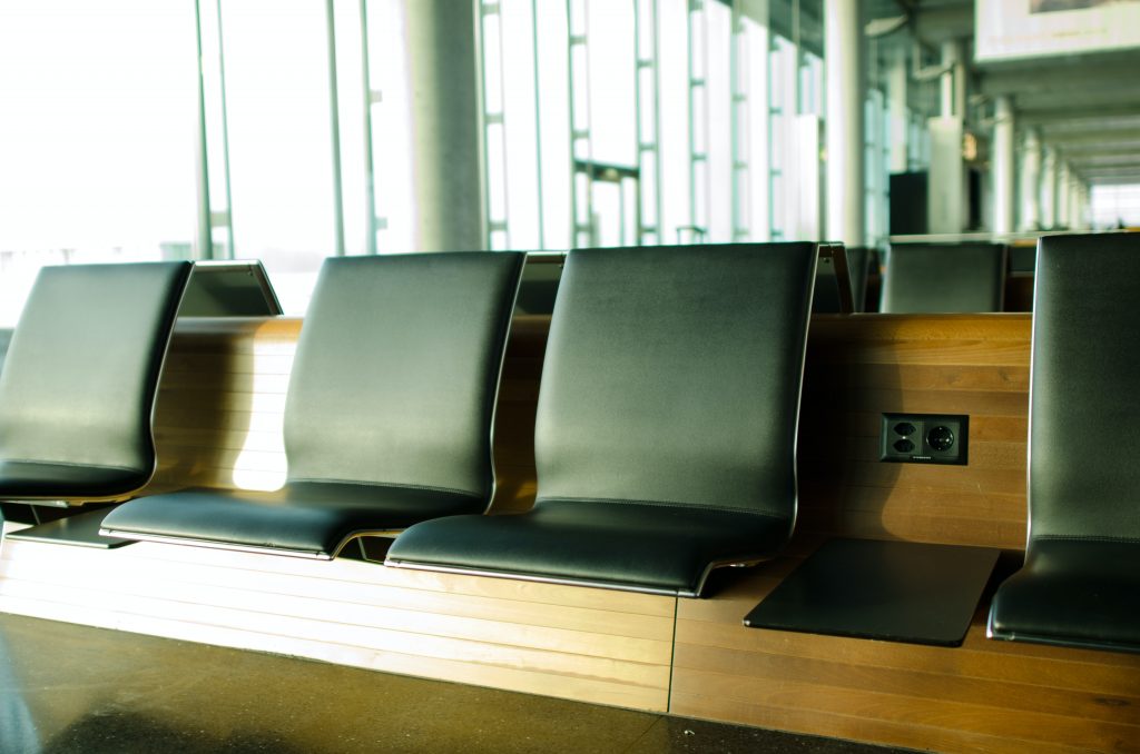 Na imagem há a representação de uma sala de check-in em aeroporto, com bancos em couro preto sobre uma estrutura de madeira.